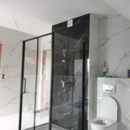 Nowoczesna kabina prysznicowa z czarnymi ramami i deszczownicą, zamontowana w łazience wykończonej marmurem, z widoczną toaletą podwieszaną i niedokończonymi elementami instalacji elektrycznej.