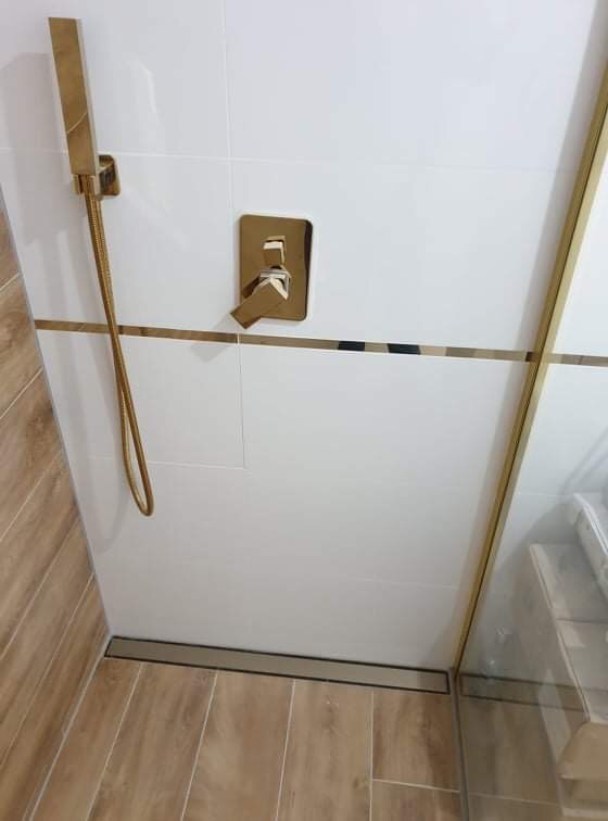 Nowoczesna kabina prysznicowa z białymi płytkami, złotymi akcentami i drewnopodobną podłogą, widoczny odpływ liniowy.