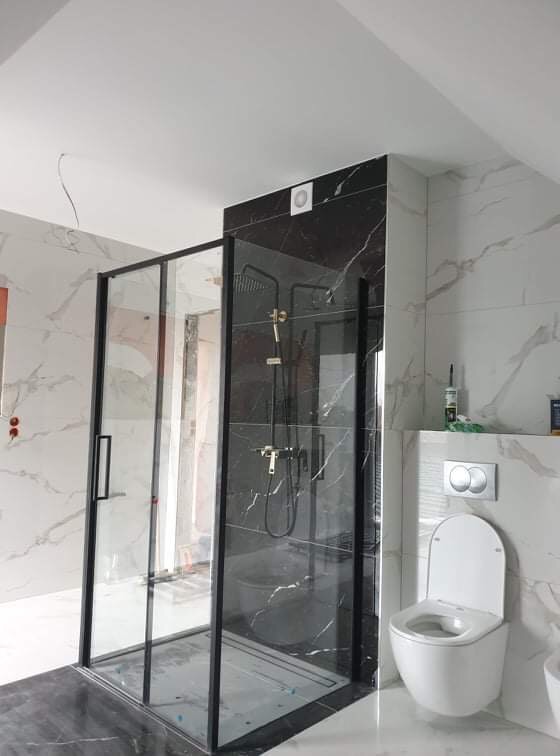 Nowoczesna kabina prysznicowa z czarnymi ramami i deszczownicą, zamontowana w łazience wykończonej marmurem, z widoczną toaletą podwieszaną i niedokończonymi elementami instalacji elektrycznej.