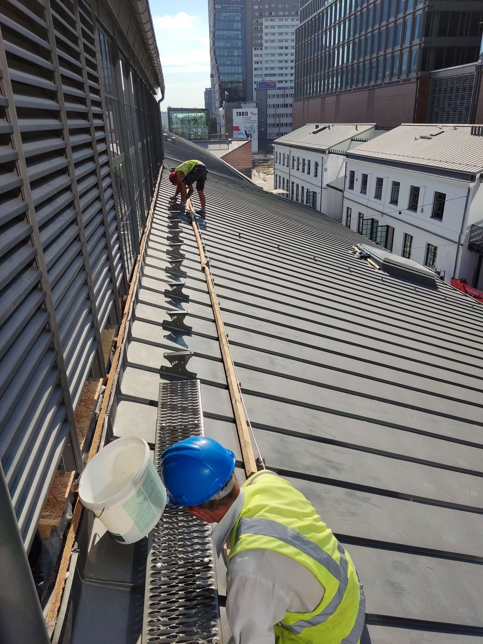 Pracownicy w odblaskowych kamizelkach montują system odprowadzania wody na dachu budynku w otoczeniu nowoczesnej architektury miejskiej.
