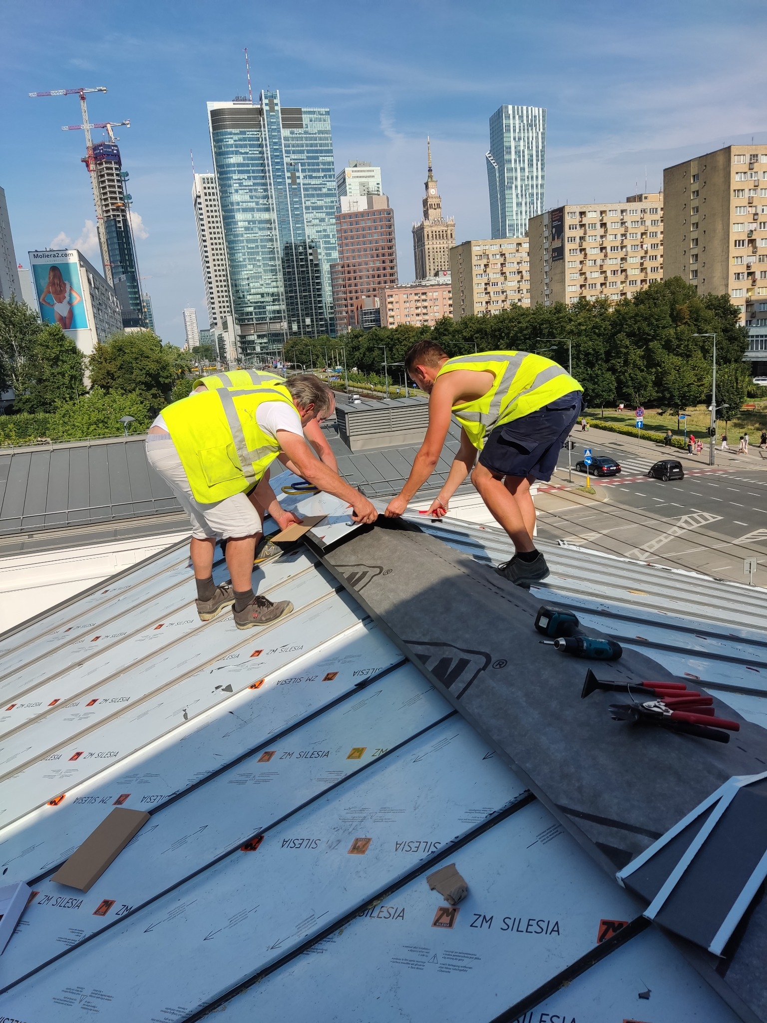 Dwóch dekarzy w żółtych kamizelkach montuje czarną membranę na metalowym dachu z widokiem na panoramę Warszawy.