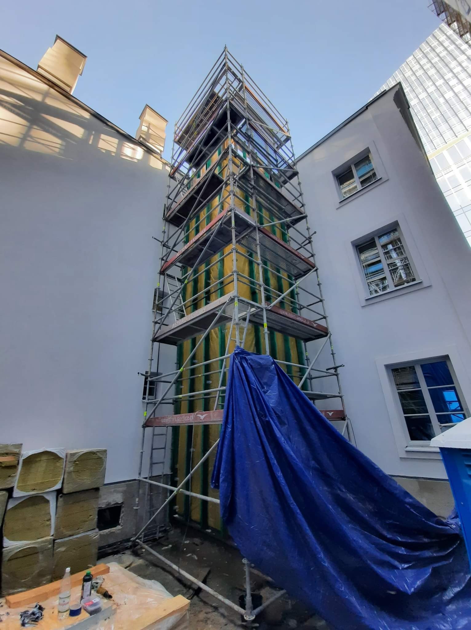 Elewacja budynku w trakcie ocieplania, widoczne rusztowania, izolacja termiczna w żółto-zielone pasy, niebieska plandeka i stos wełny mineralnej