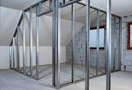 Metalowa konstrukcja szkieletowa w pomieszczeniu na poddaszu, przygotowana do montażu płyt. Widoczne skośne ściany i sufit. Częściowo otynkowane ściany z cegły. Okno w drewnianej ramie.