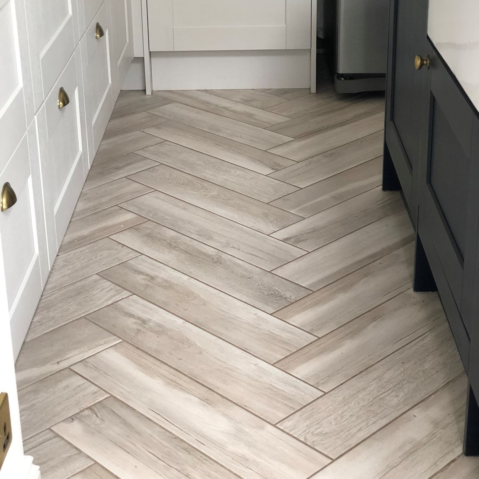 Jasne, drewnopodobne płytki ułożone w jodełkę na podłodze w kuchni, widoczne białe szafki z mosiężnymi uchwytami i fragment ciemnej zabudowy kuchennej.