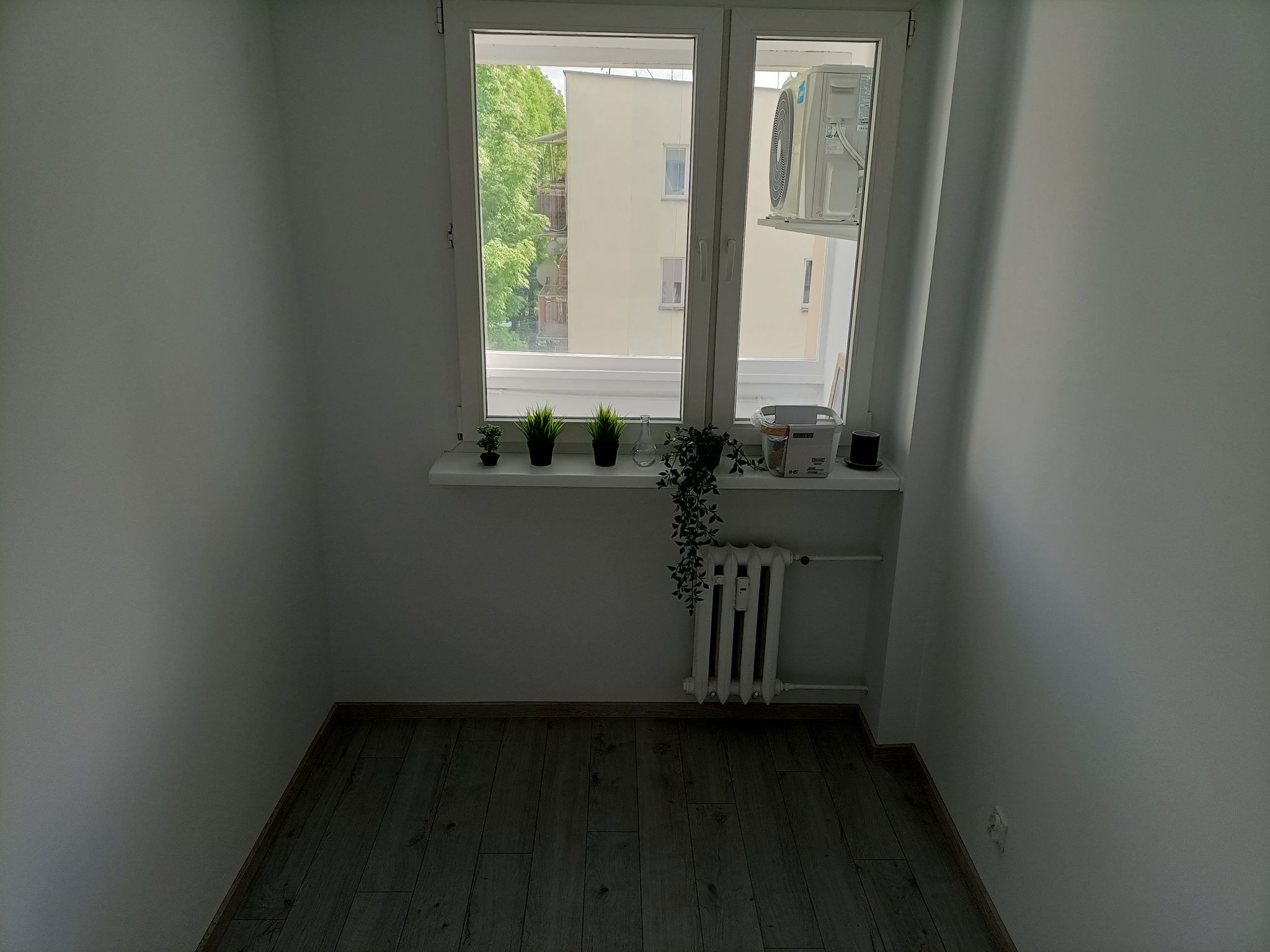 Pusty pokój po remoncie z drewnopodobną podłogą, białymi ścianami, grzejnikiem pod oknem i klimatyzatorem na zewnątrz okna.