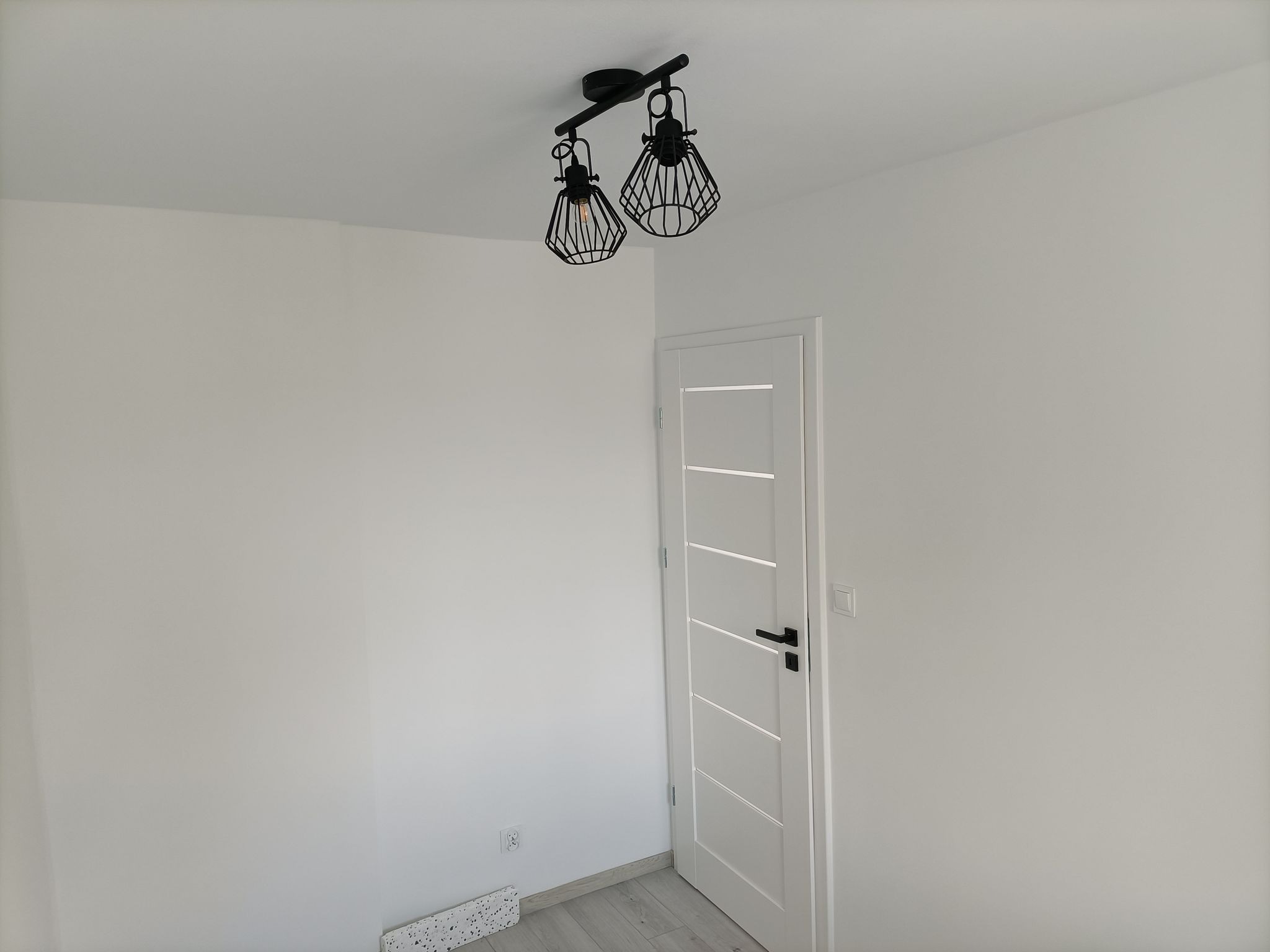 Wnętrze pomalowanego na biało pokoju z białą stolarką drzwiową i czarną lampą sufitową w stylu industrialnym.