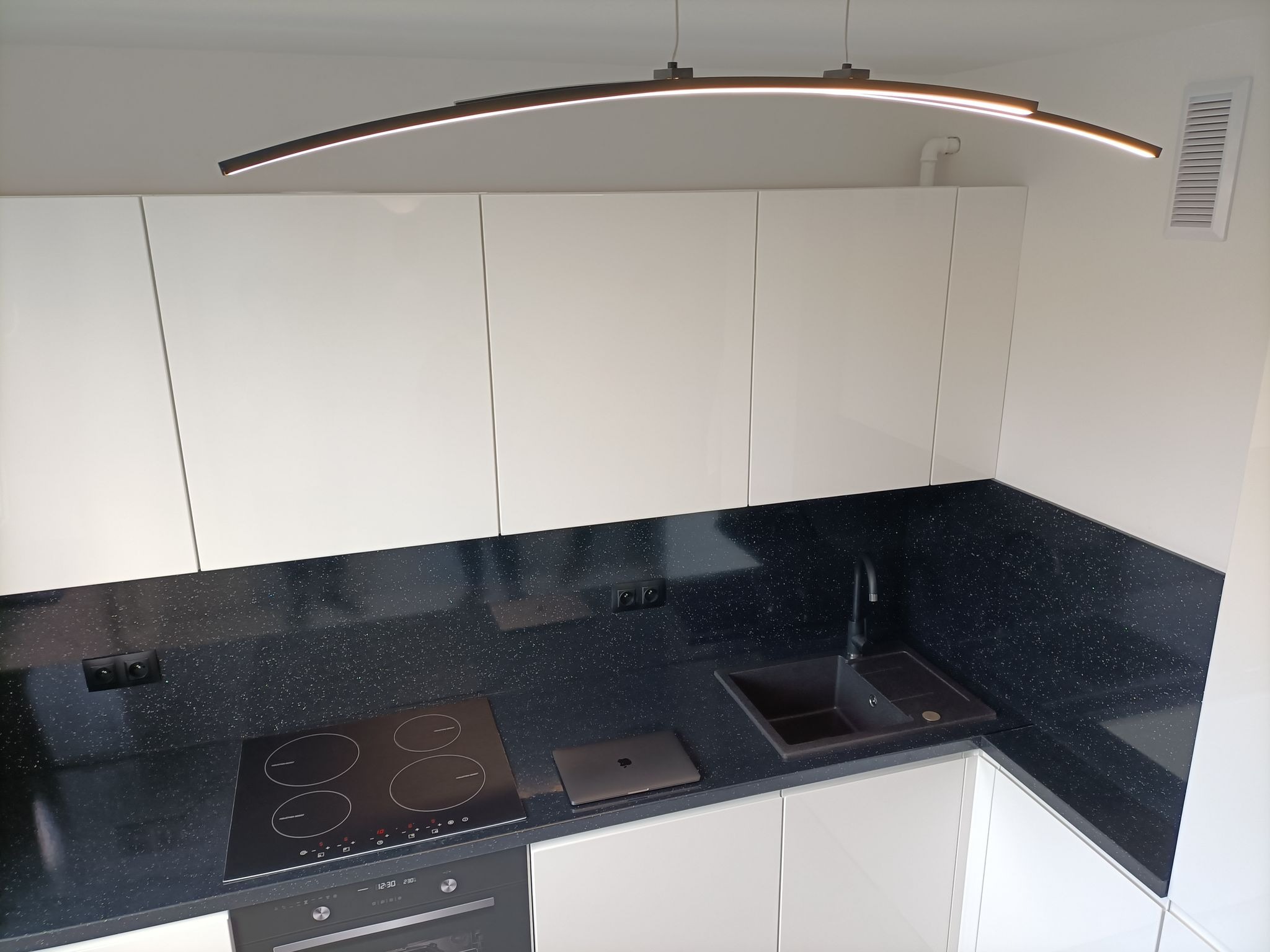 Nowoczesna kuchnia z białymi szafkami, czarnym blatem z drobinkami, indukcyjną płytą grzewczą, minimalistyczną lampą LED i laptopem na blacie.