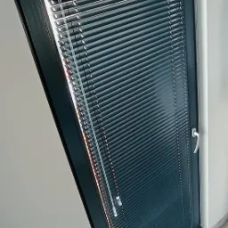 Ciemne, metaliczne żaluzje poziome zamontowane w oknie o grafitowej ramie, widok z boku.