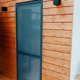 Drewniana elewacja budynku z zamontowaną moskitierą w ciemnej ramie, oświetlona dwoma czarnymi kinkietami.