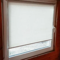 Beżowa tekstylna osłona okienna częściowo zwinięta w oknie z białego PCV w domu z boazerią.