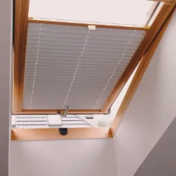 Okno dachowe z drewnianą ramą i zamontowaną plisą w kolorze jasnoszarym, widok na dach sąsiedniego budynku.