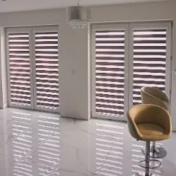 Eleganckie wnętrze z dwoma oknami przesłoniętymi roletami typu dzień i noc w kolorze brązowo-białym, marmurowa podłoga odbijająca światło, designerski żyrandol i dwa beżowe krzesła barowe.