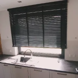 Ciemne poziome osłony okienne zamontowane w kuchni nad zlewem, częściowo zasłonięte, z widocznymi elementami mocującymi.
