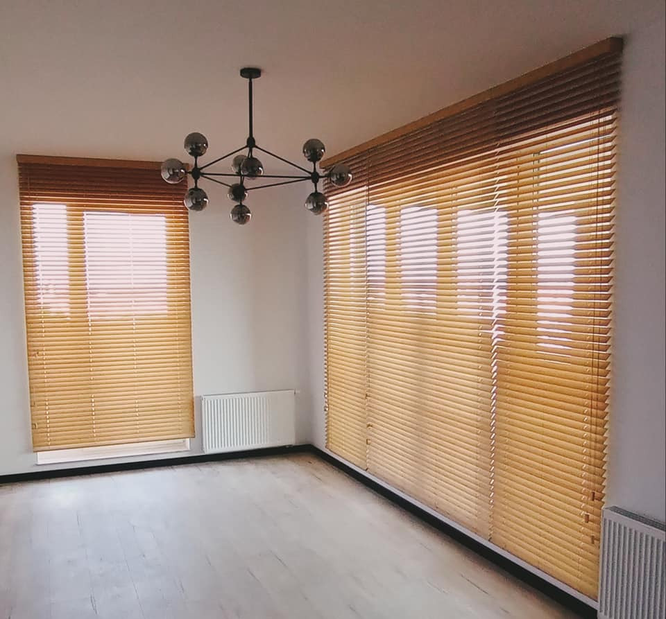 Jasne drewniane żaluzje poziome w pustym pokoju z nowoczesnym żyrandolem, widok na okna i grzejniki.