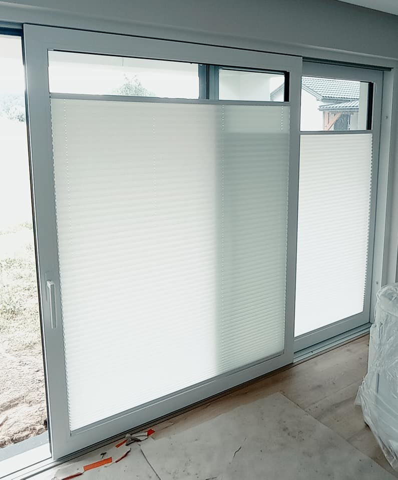 Białe, plisowane osłony okienne zamontowane w nowoczesnych, przesuwnych oknach balkonowych z szarymi ramami, widoczne niedokończone prace wykończeniowe podłogi.