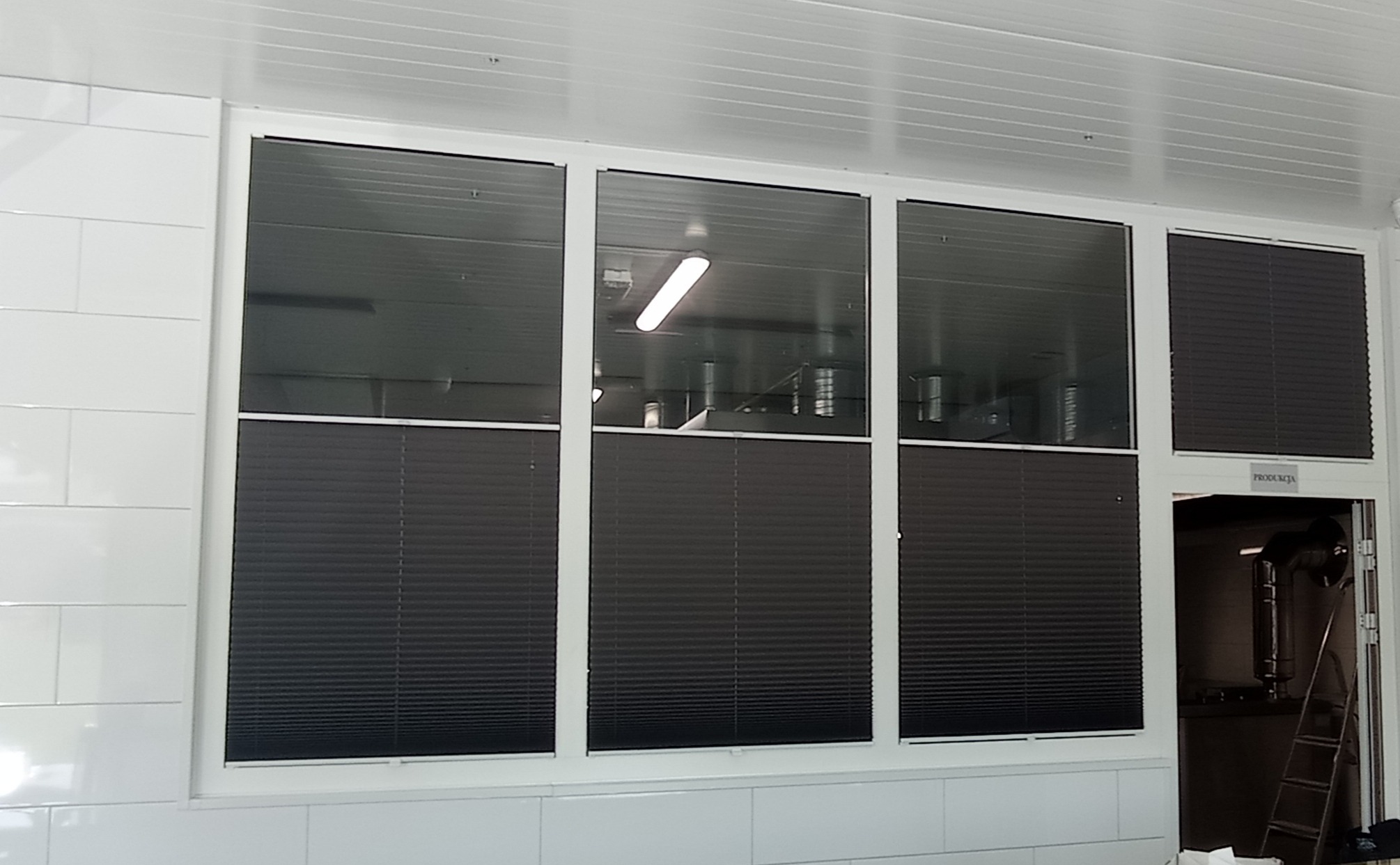 Ciemne plisy okienne zamontowane w oknach pomieszczenia produkcyjnego z białymi, kafelkowymi ścianami i sufitem panelowym.