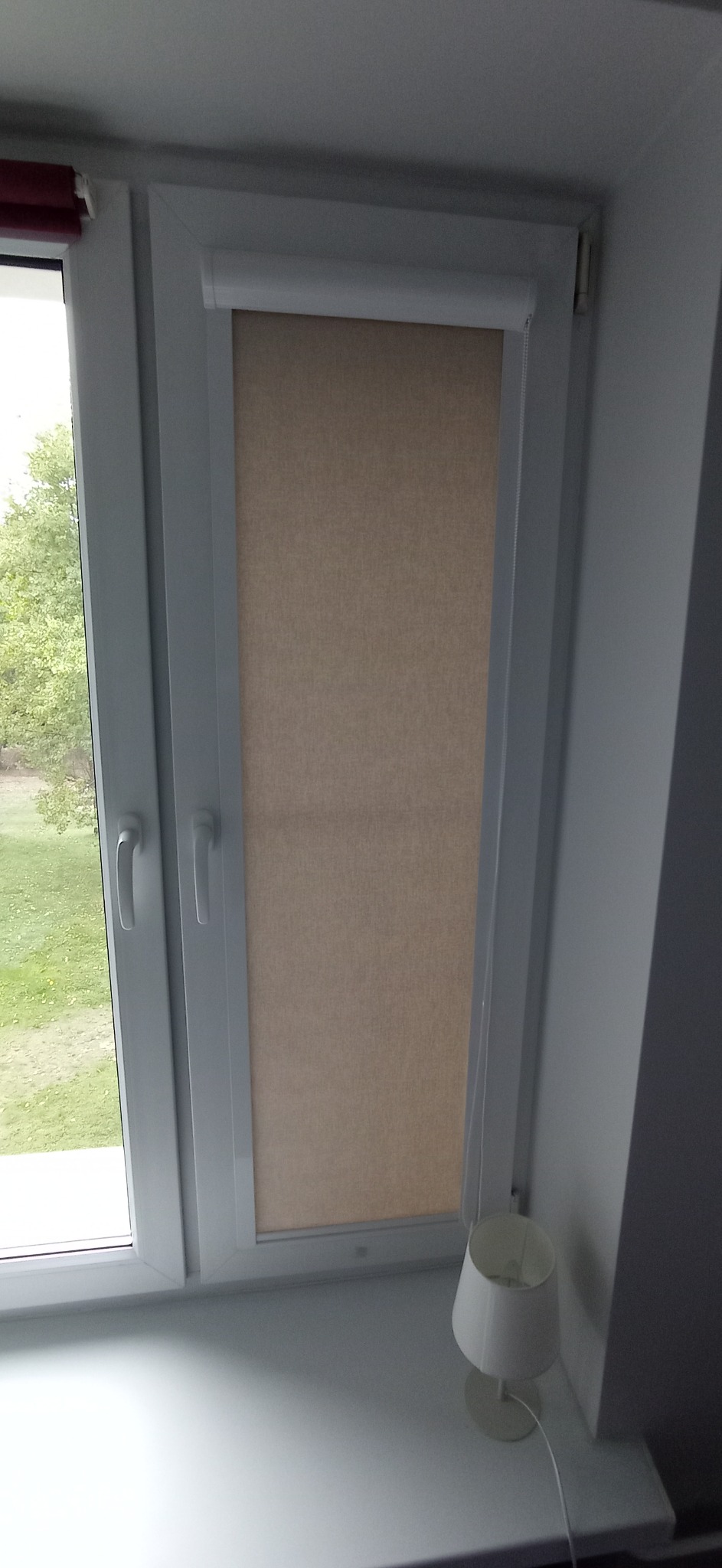 Zamknięta, beżowa osłona okienna w białej ramie okna, widok z wnętrza pomieszczenia, obok lampka nocna.