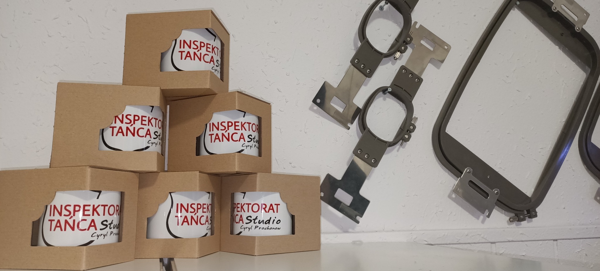Stos kubków w brązowych pudełkach z nadrukiem logo 'Inspektor Tańca Studio' oraz metalowe ramki do haftowania na ścianie.