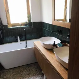 Nowoczesna łazienka z wanną, dwiema umywalkami nablatowymi na drewnianej szafce, ciemnozielonymi płytkami na ścianie i oknem z drewnianą ramą.