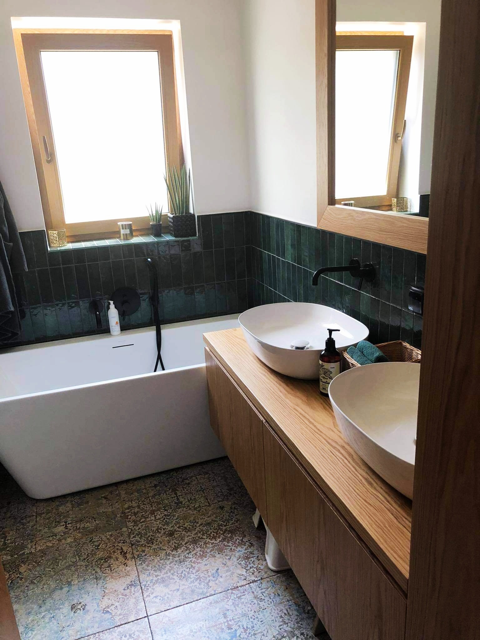 Nowoczesna łazienka z wanną, dwiema umywalkami nablatowymi na drewnianej szafce, ciemnozielonymi płytkami na ścianie i oknem z drewnianą ramą.