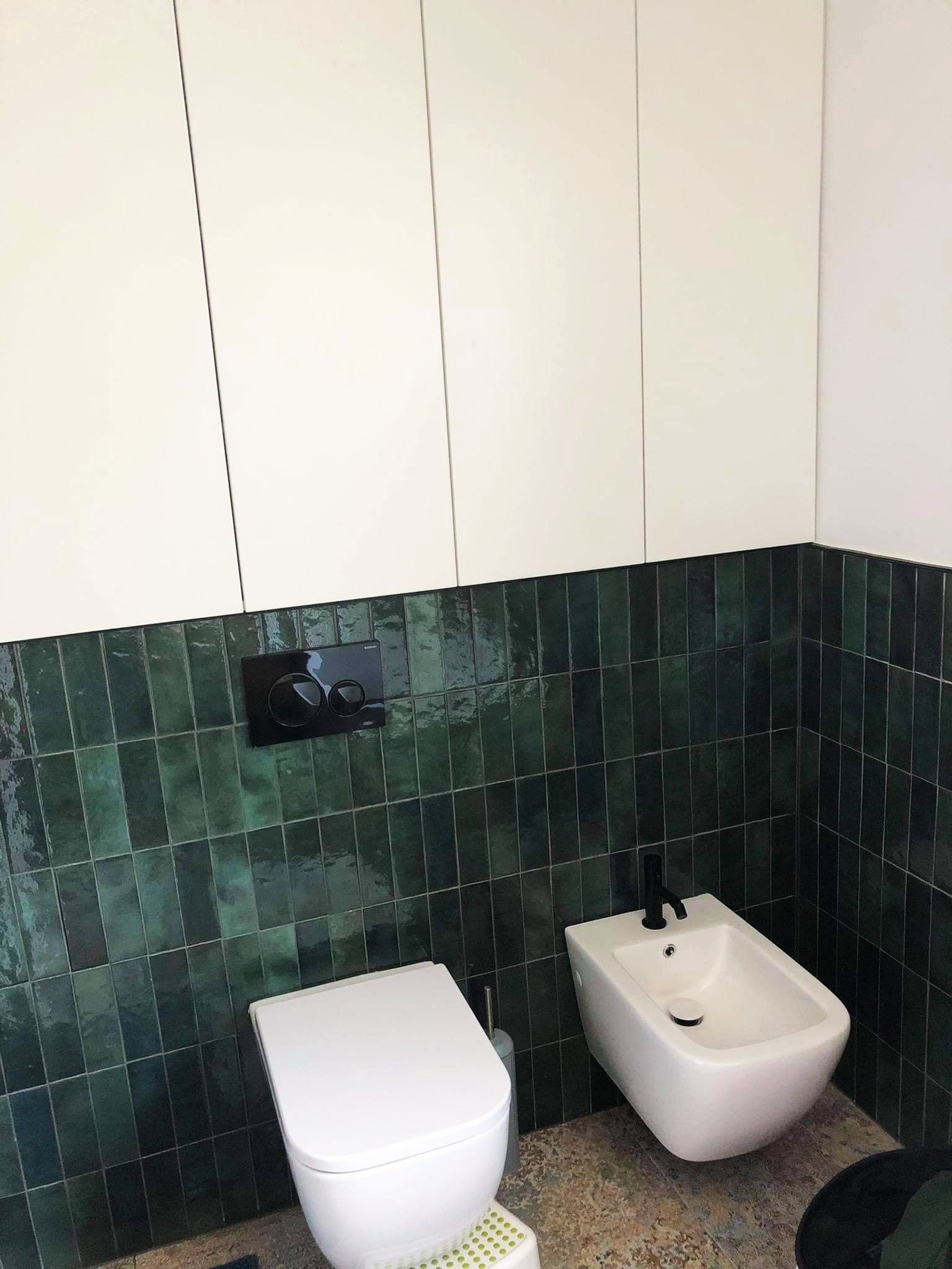 Nowoczesna łazienka z białą toaletą i bidetem z czarną armaturą, zielone, wąskie płytki na ścianie, białe szafki wiszące, widoczny podest dla dziecka.