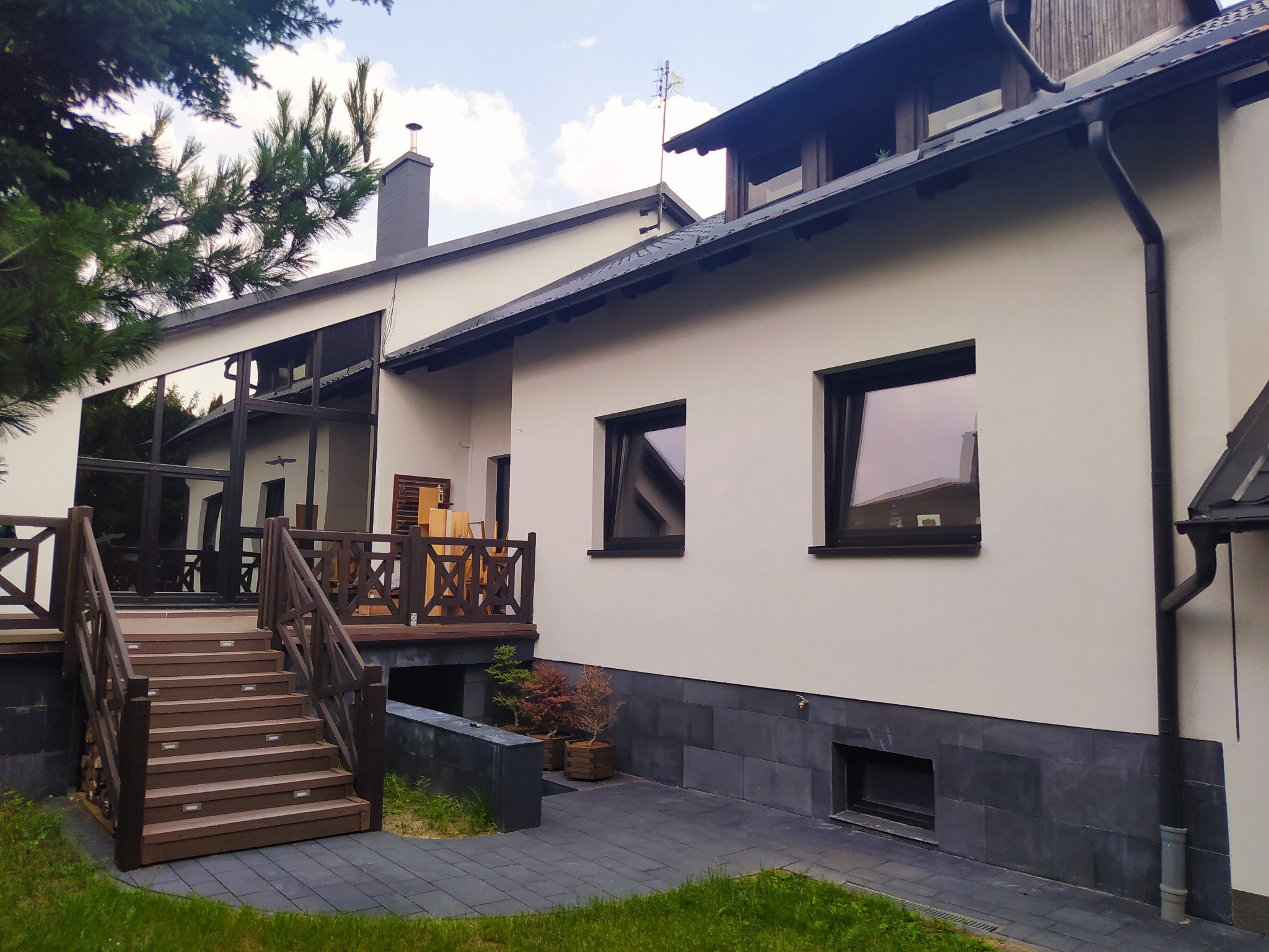 Elewacja domu z drewnianym tarasem i schodami, widoczne okna z czarnymi ramami, szara podmurówka i fragment ogrodu z drzewami iglastymi.