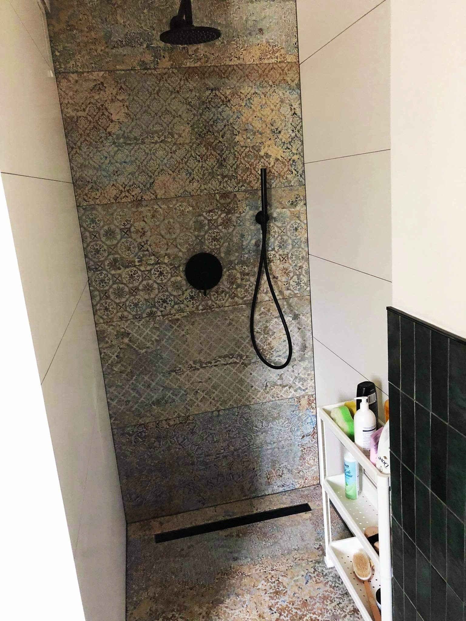 Wykończona kabina prysznicowa z czarną armaturą i odpływem liniowym, ściany wyłożone płytkami w stylu vintage, obok biały regał z kosmetykami.