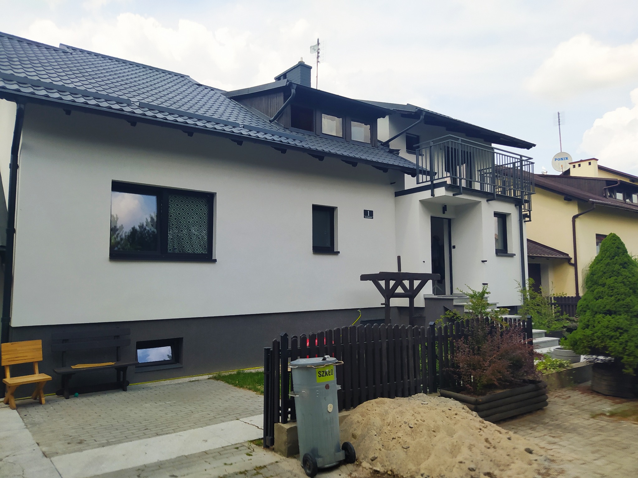 Biały dom z ciemną dachówką i balkonem, widoczny z zewnątrz, z elementami krajobrazu, takimi jak ogrodzenie, drzewa i trawnik, oraz śmietnikiem na pierwszym planie.