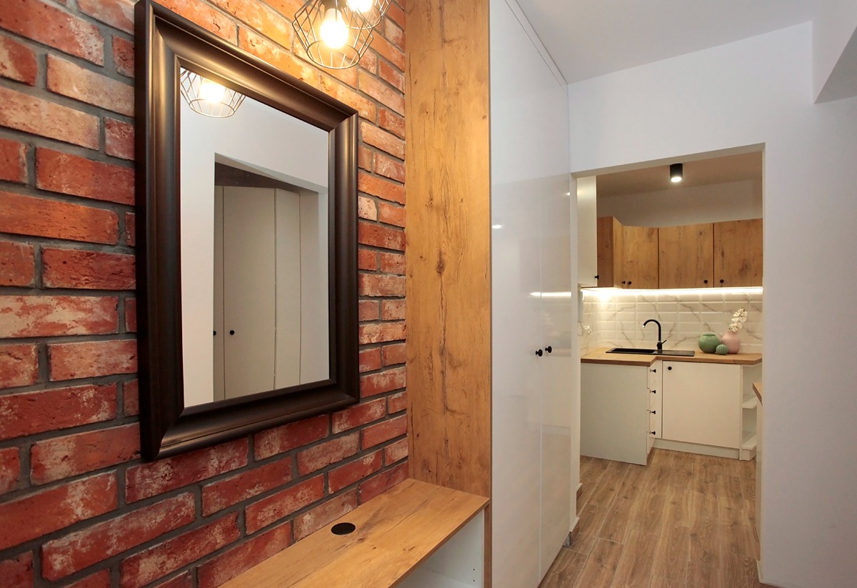 Ceglana ściana z lustrem w ciemnej ramie i lampą drucianą, widok na odnowioną kuchnię z białymi szafkami i drewnianym blatem, podłoga imitująca drewno.