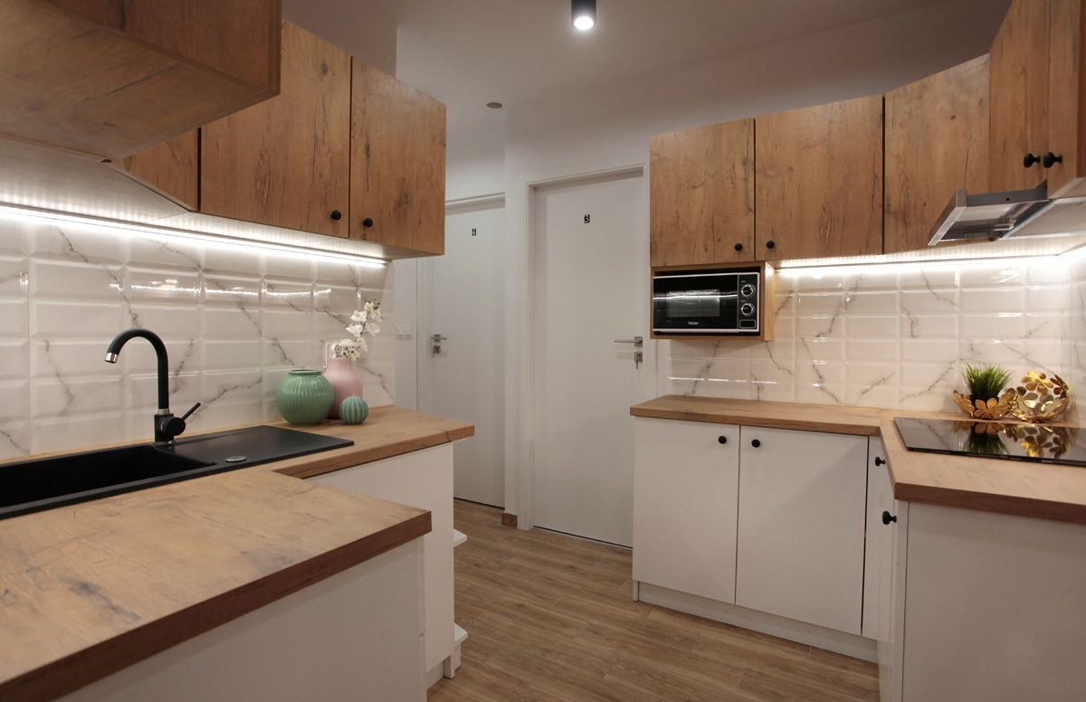 Jasna kuchnia z drewnianymi szafkami górnymi, białymi szafkami dolnymi, czarnym zlewem i armaturą, oraz białymi, marmurkowymi płytkami na ścianie, oświetlona listwą LED pod szafkami.