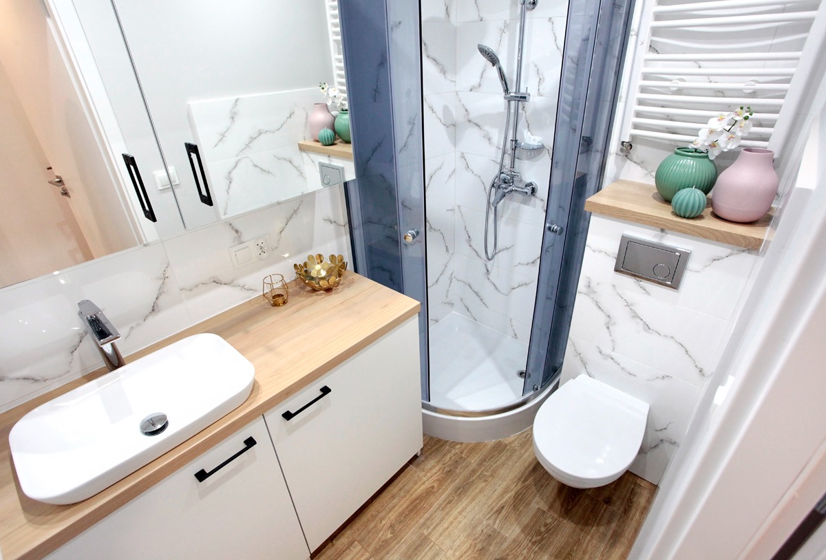 Nowoczesna łazienka z kabiną prysznicową w szarej ramie, umywalką nablatową na drewnianym blacie, białymi szafkami z czarnymi uchwytami i marmurkowymi płytkami na ścianach.