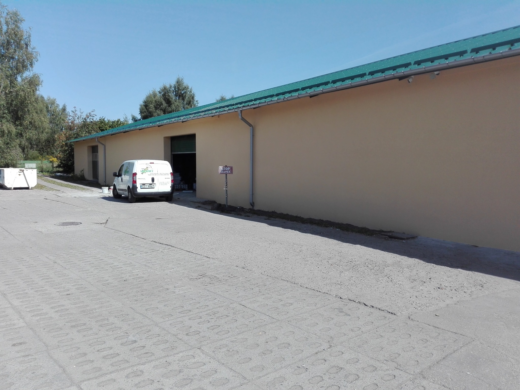 Budynek magazynowy z zielonym dachem i kremową elewacją, z zaparkowanym białym busem z logo firmy przed otwartą bramą. Na pierwszym planie betonowy plac z wzorem kostki.