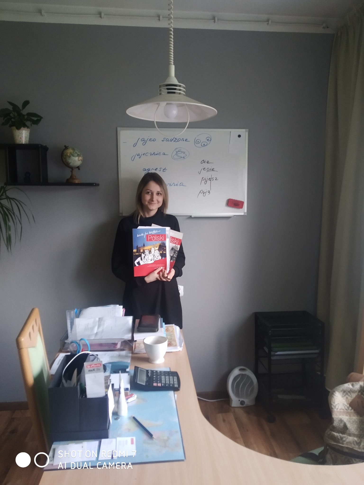 Kobieta trzymająca podręczniki do nauki języka polskiego na tle tablicy z napisanymi słowami i wyrażeniami po polsku w domowym gabinecie.