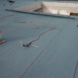 Niebieska papa termozgrzewalna na płaskim dachu, widoczny palnik dekarski z płomieniem i butla gazowa, w tle surowy budynek w trakcie budowy.