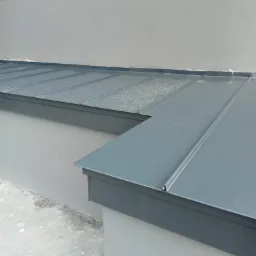 Szary dach z blachy trapezowej, połączony z obróbką blacharską przy ścianie budynku, widoczne ślady wilgoci na powierzchni.