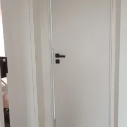 Białe drzwi wewnętrzne z czarną klamką w minimalistycznym wnętrzu. Podłoga z paneli imitujących drewno, widoczny fragment futryny drzwiowej.