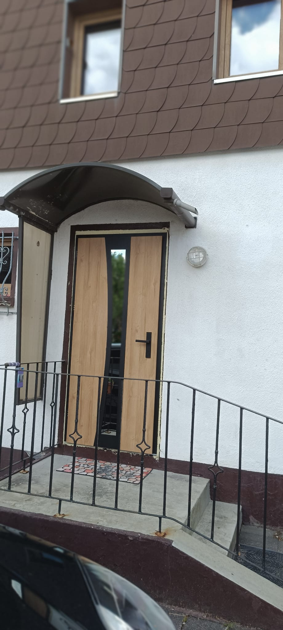 Zewnętrzne drzwi wejściowe z drewnianym panelem i czarnymi wstawkami, osłonięte daszkiem. Widoczne schody z metalową barierką i fragment elewacji budynku.