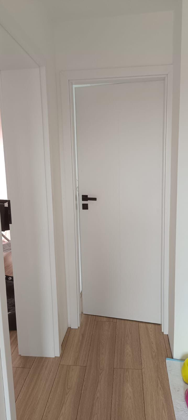 Białe drzwi wewnętrzne z czarną klamką w minimalistycznym wnętrzu. Podłoga z paneli imitujących drewno, widoczny fragment futryny drzwiowej.