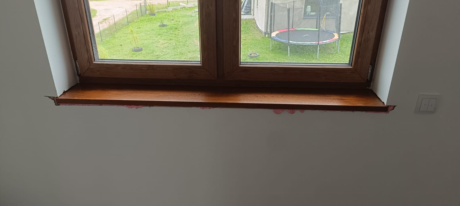 Drewniane okno z widokiem na zewnątrz, z widocznymi śladami pianki montażowej na ścianie i parapet z połyskliwą powierzchnią. W tle zielony trawnik i trampolina.