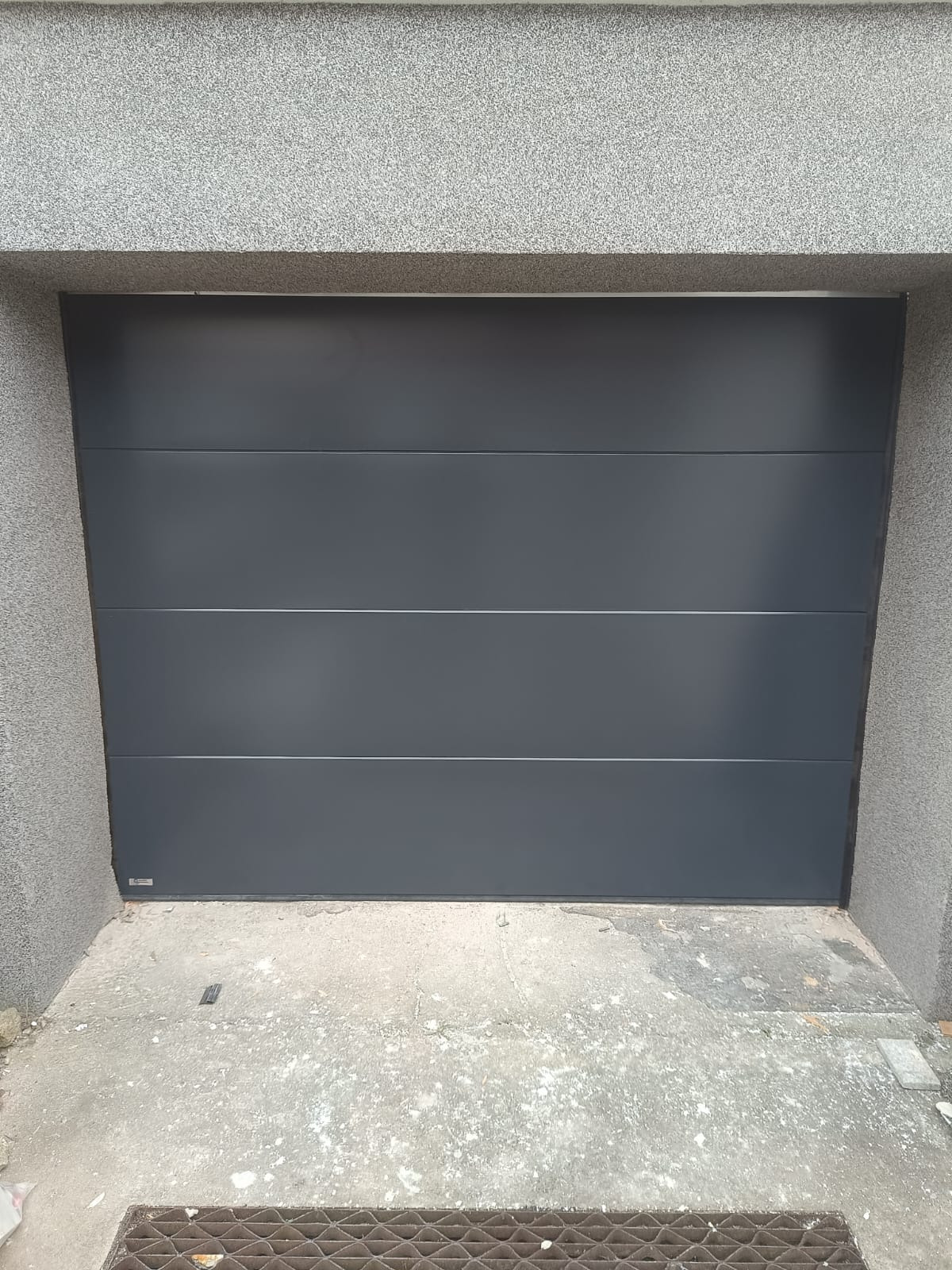Zamknięta, segmentowa brama garażowa w odcieniach szarości, z widoczną fakturą ściany garażu i betonową posadzką. Minimalistyczny design, równoległe panele.