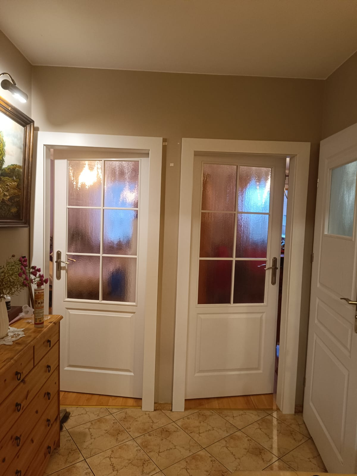 Dwa białe, przeszklone skrzydła drzwiowe wewnętrzne, zamontowane w ościeżnicach. Widoczna podłoga z płytek i fragment komody. Neutralne tło ścian.