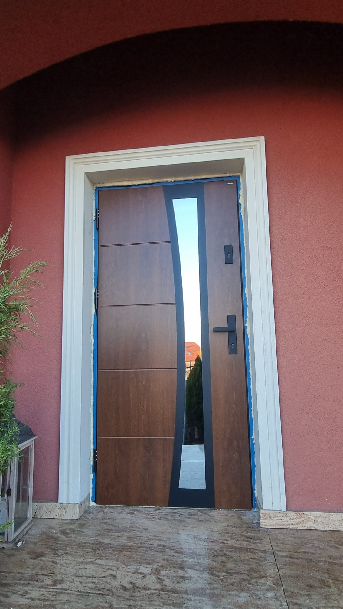 Nowoczesne drzwi zewnętrzne z przeszkleniem, brązowe, zamontowane w białej futrynie na tle czerwonej ściany. Widoczne zabezpieczenia montażowe.