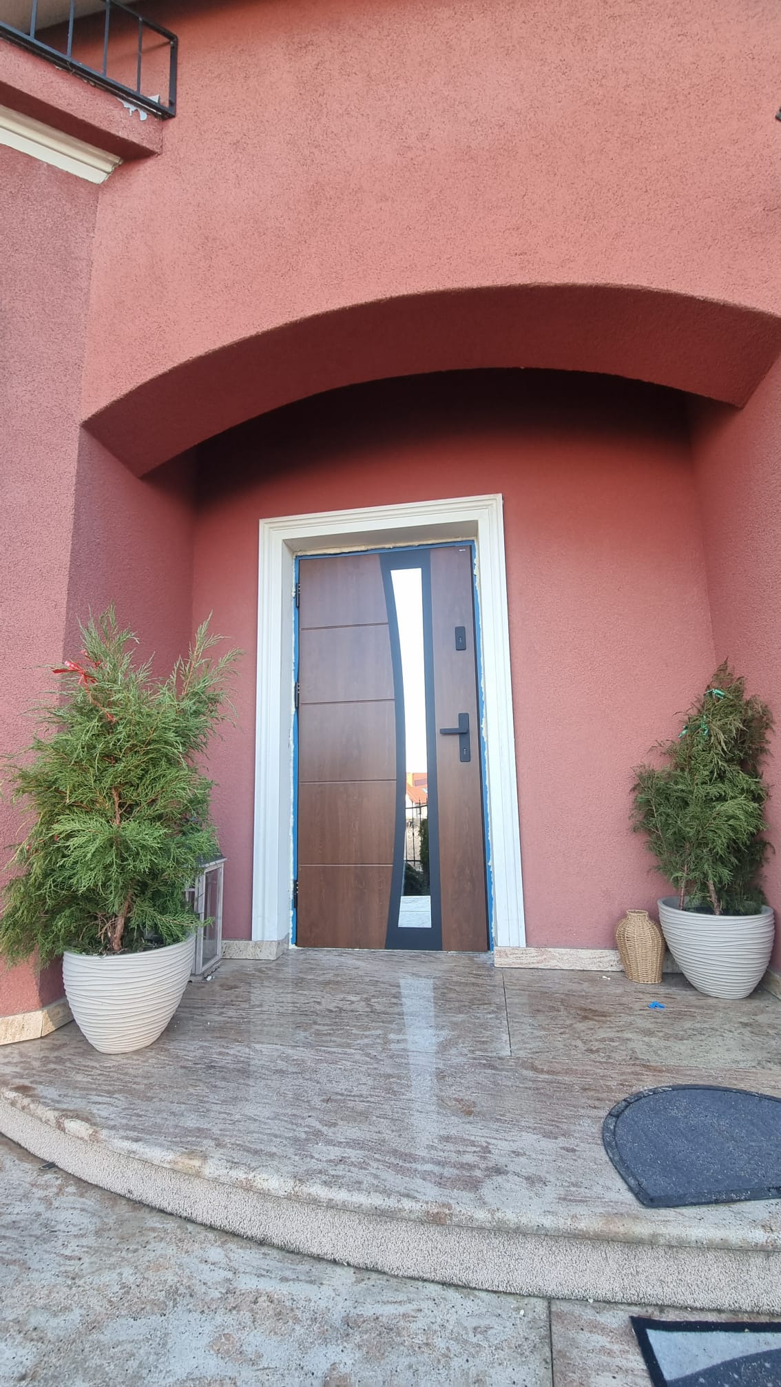 Brązowe drzwi zewnętrzne z przeszkleniem, osadzone w białej futrynie, na tle różowej elewacji z łukiem. Po bokach drzwi stoją tuje w białych donicach.