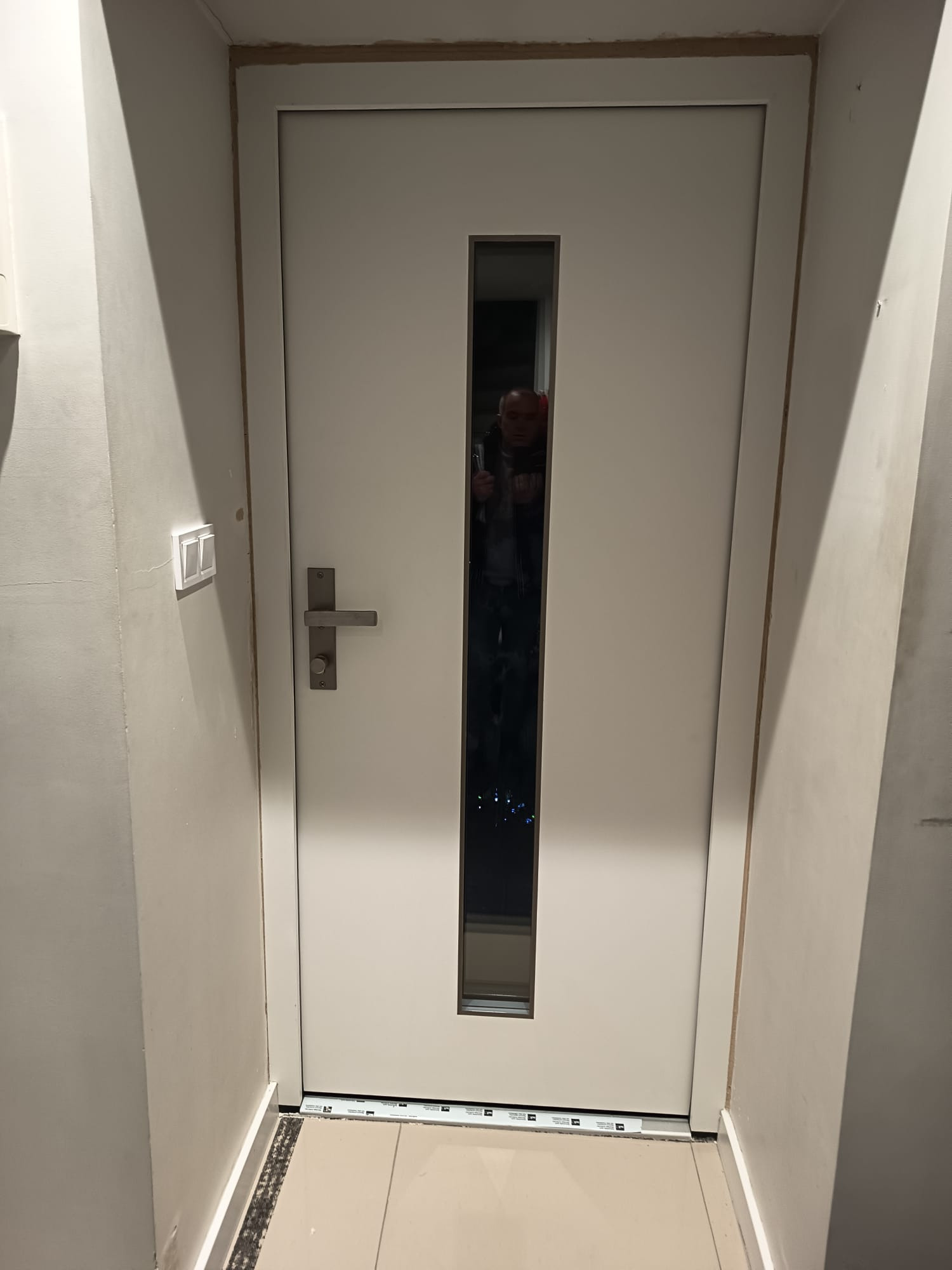 Białe drzwi wewnętrzne z wąskim przeszkleniem, widoczny fragment ościeżnicy i podłogi. W odbiciu szyby widoczna osoba robiąca zdjęcie.