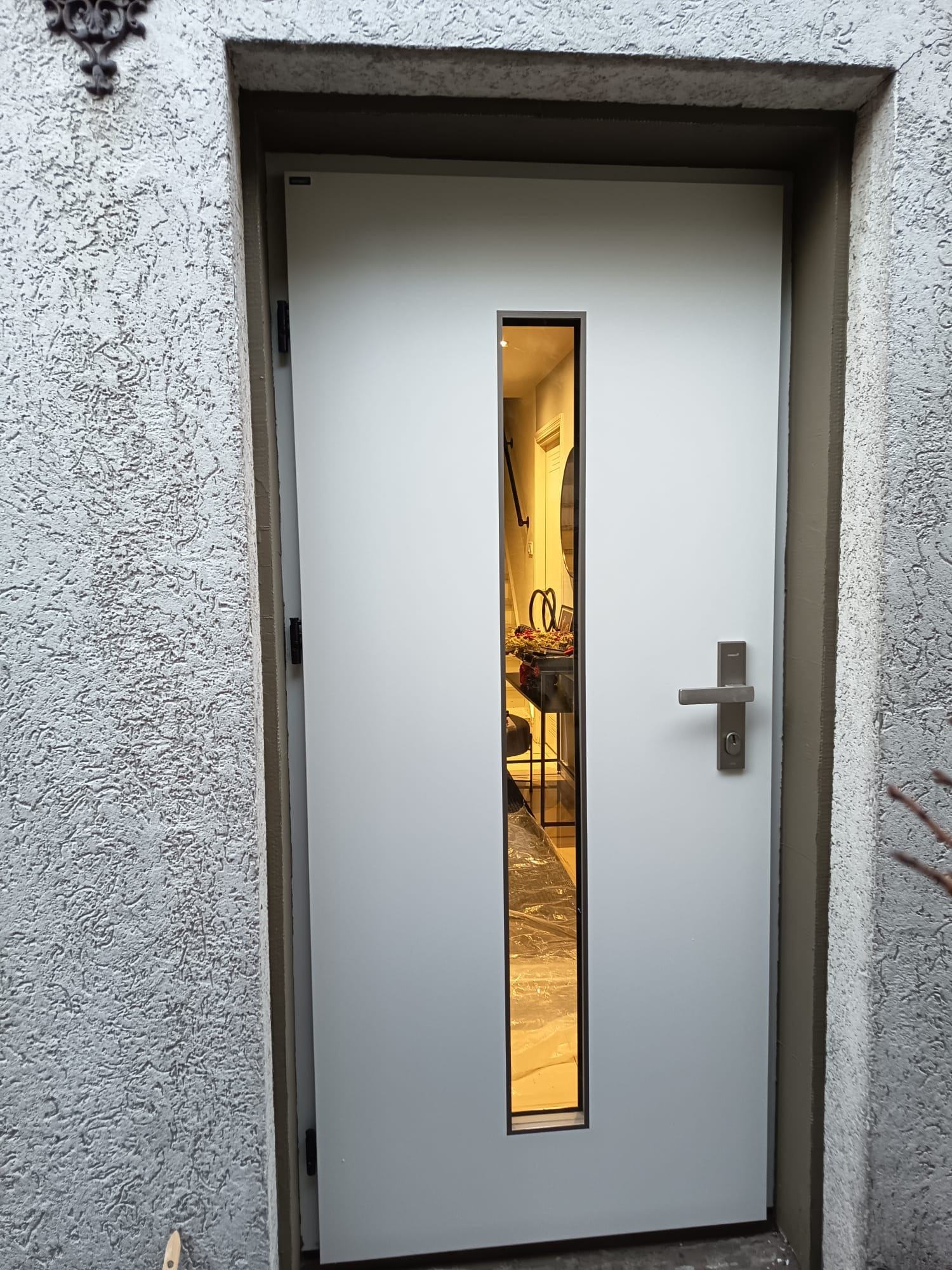 Białe drzwi zewnętrzne z wąskim, pionowym przeszkleniem, widok na wnętrze domu. Szara, chropowata elewacja budynku. Metalowa klamka i zamek.