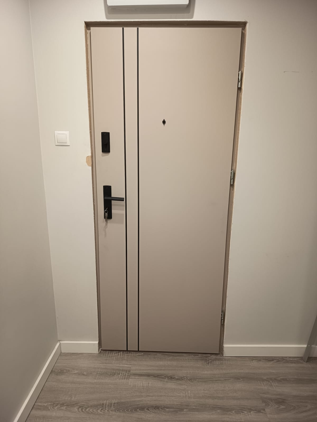 Beżowe drzwi z czarnymi, pionowymi paskami i czarną klamką na tle szarej ściany i podłogi imitującej drewno. Widoczne zawiasy i wizjer.