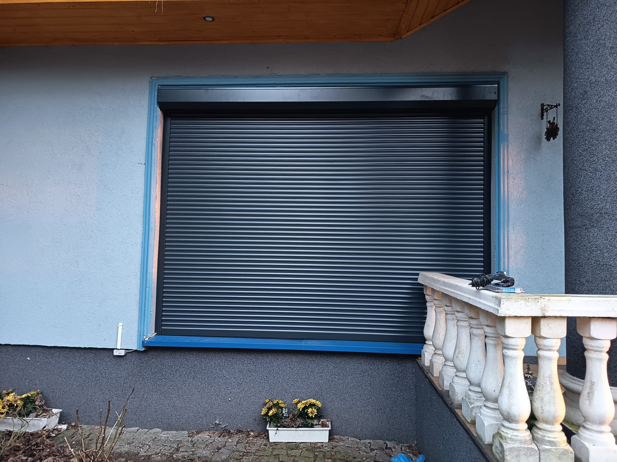 Zamknięta, czarna roleta zewnętrzna na tle błękitnej ściany domu z dekoracyjnym obramowaniem okna. Widoczne kwiaty w doniczce i fragment balustrady.