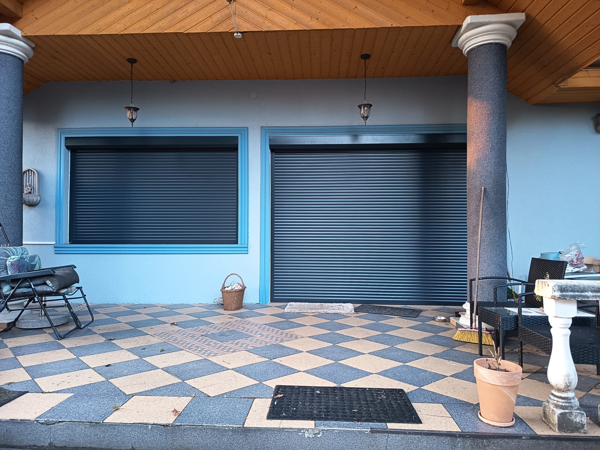 Błękitny budynek z dwoma oknami i bramą garażową z roletami zewnętrznymi, kolumnami i szachownicową podłogą.