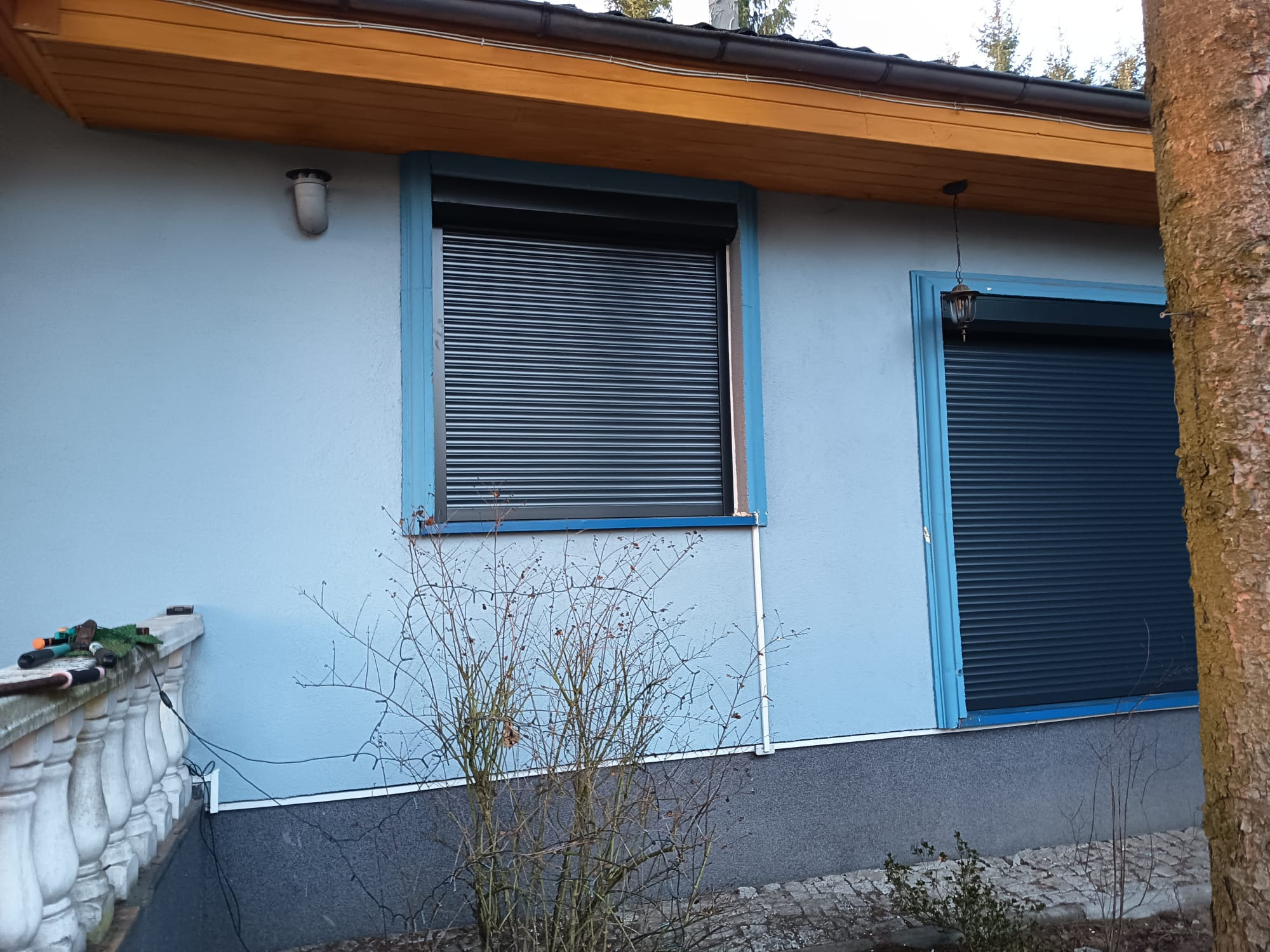Niebieski dom z zamontowanymi, zamkniętymi roletami zewnętrznymi w kolorze grafitowym. Widoczne narzędzia ogrodowe na balustradzie i fragment pnia drzewa.