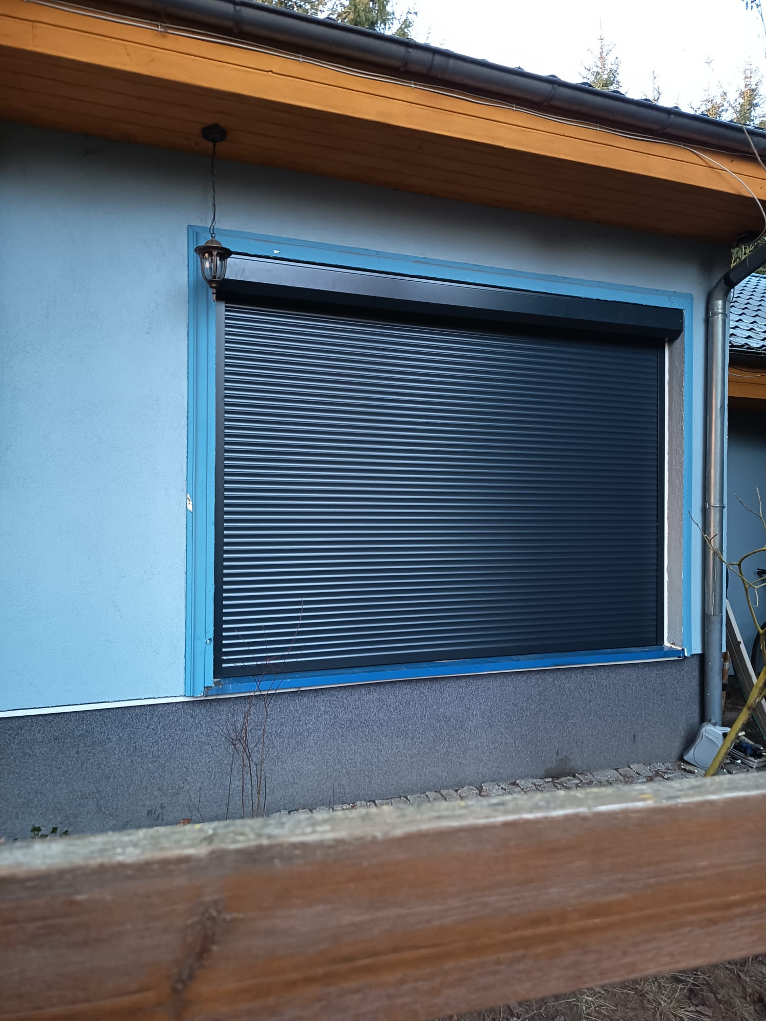 Zewnętrzna roleta okienna w kolorze grafitowym, zamontowana w niebieskiej elewacji budynku. Widoczny fragment drewnianego płotu i lampa zewnętrzna.